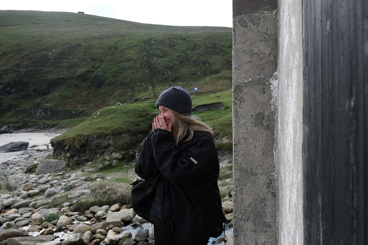 Caroline Wosskow smiling into her hands on a rocky coastline beside a concrete pillar