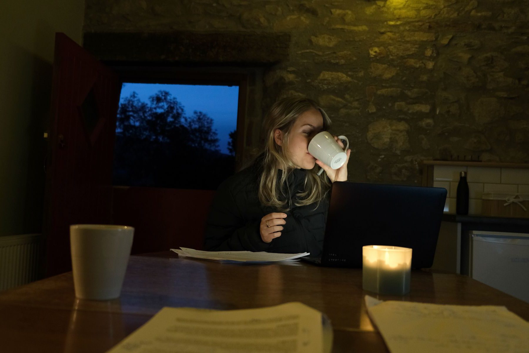Caroline Wosskow drinking from a mug while writing at night by candlelight, scripts on the table in front of her